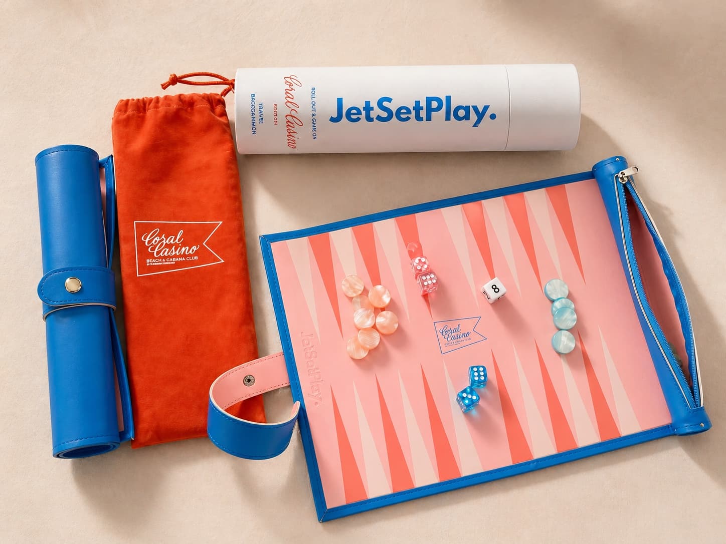 Coral Casino travel backgammon set on warm cream: a blue leather roll, an orange pouch with a script flag mark, a white JetSetPlay tube, and the open board with coral points, pearlescent chips and dice